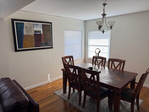 A dining area with a wooden table and chairs, a chandelier, a framed abstract painting, and a window with blinds. - Cozy 2-bedroom house in peaceful Corpus Christi close to hospital beach downtown (Corpus Christi)