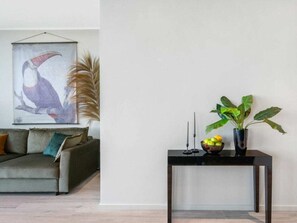 Ein modernes Wohnzimmer mit einem grauen Sofa, einem gerahmten Kunstwerk mit einem Vogel, einer hohen Pflanze und einem schwarzen Beistelltisch mit einer Obstschale.