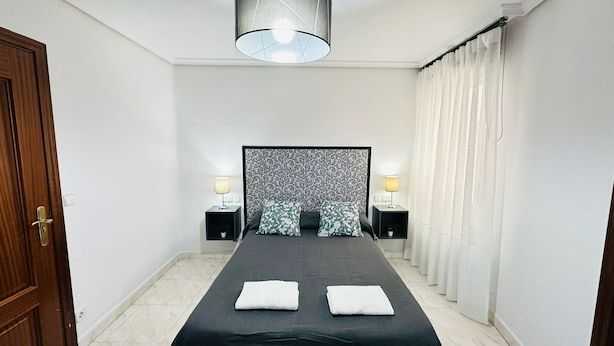 Ein Hotelzimmer mit einem Bett, zwei Nachttischlampen, einem Vorhang mit Muster und einer Tür links.