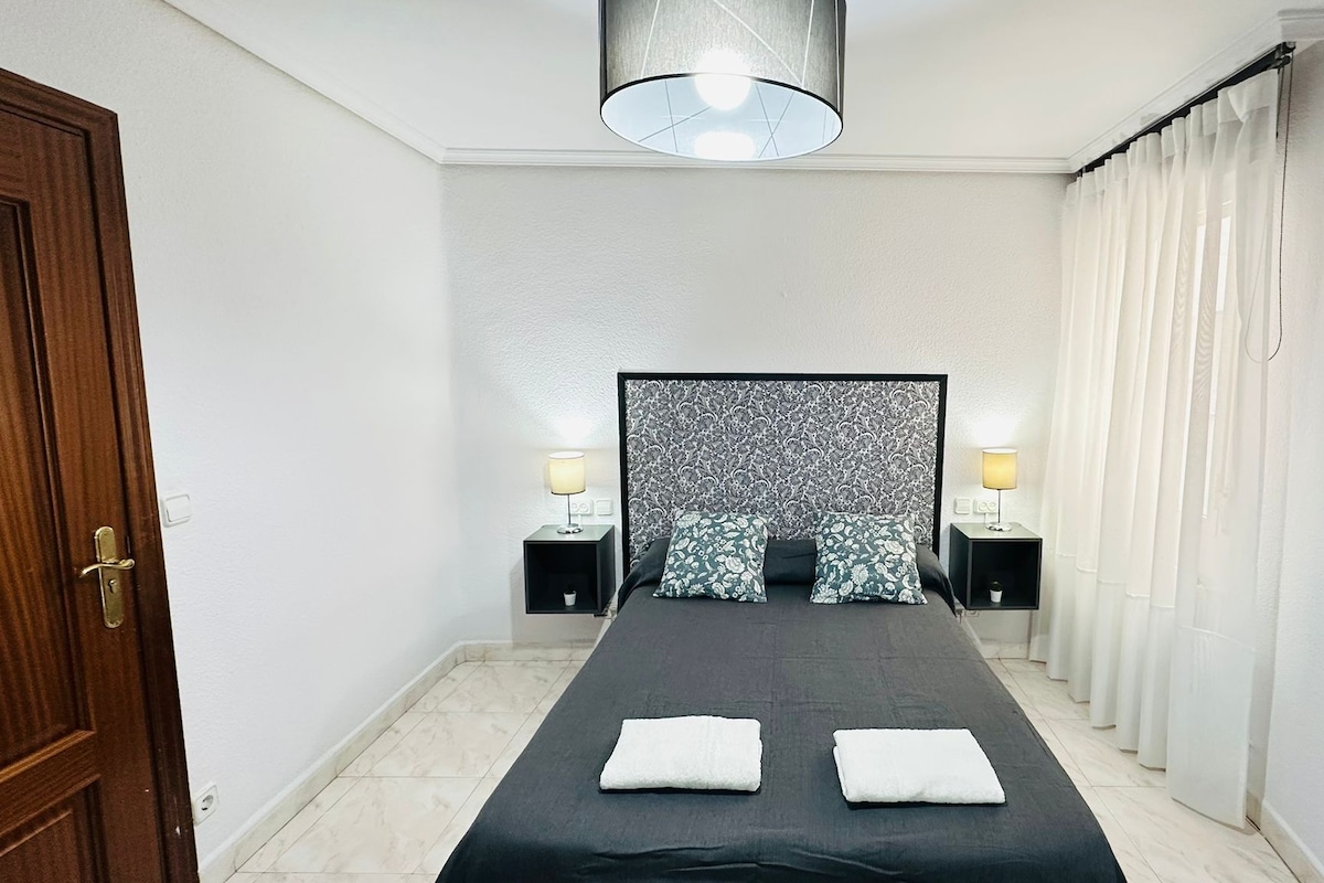 Una habitación de hotel con una cama, dos lámparas de noche, un cabecero con diseño estampado y una puerta a la izquierda.