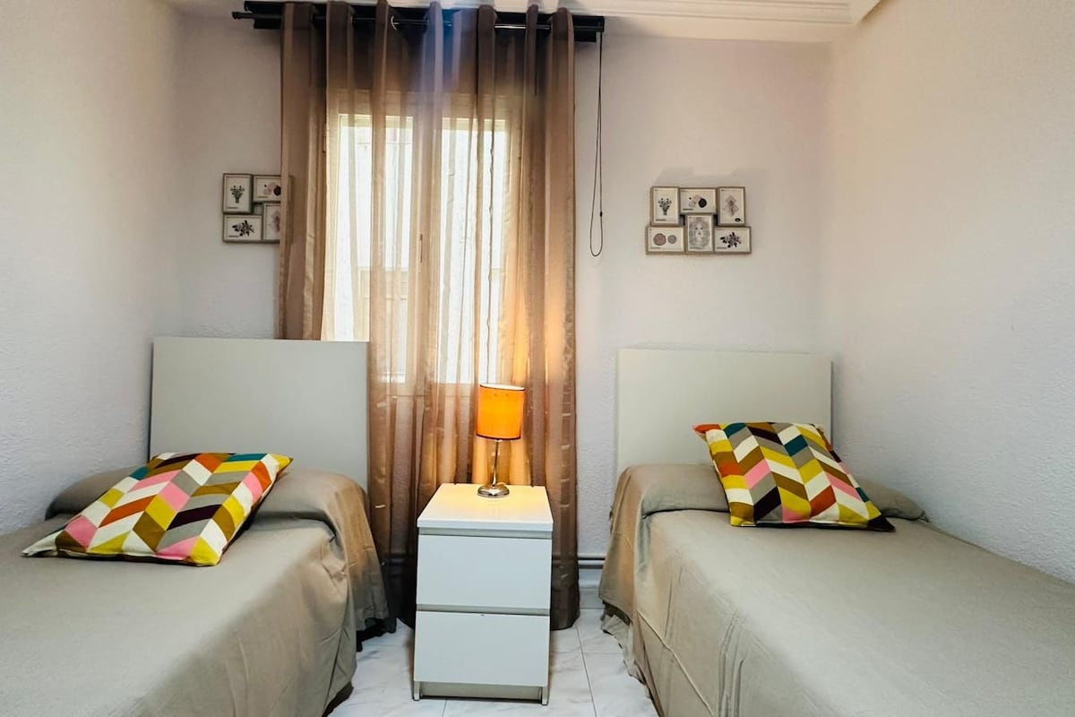 Una habitación con dos camas, una mesita de noche con lámpara y cortinas.