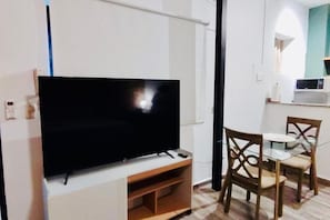 Ein Flachbildfernseher auf einem Standfuß, ein Esstisch mit Glasplatte und zwei Stühlen sowie eine Mikrowelle auf einer Arbeitsplatte.