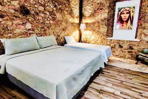 Een kamer met een stenen muur, een bed met een witte spreidek, een nachtkastje met een lamp en een ingelijst schilderij aan de muur.