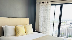 A modern bedroom with a large bed, a headboard, and a view of the city through a large window. - Apartment in Sandton (Sandton)