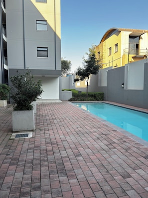 A swimming pool area with a brick pathway, a modern building, and a tree. - Apartment in Sandton (Sandton)