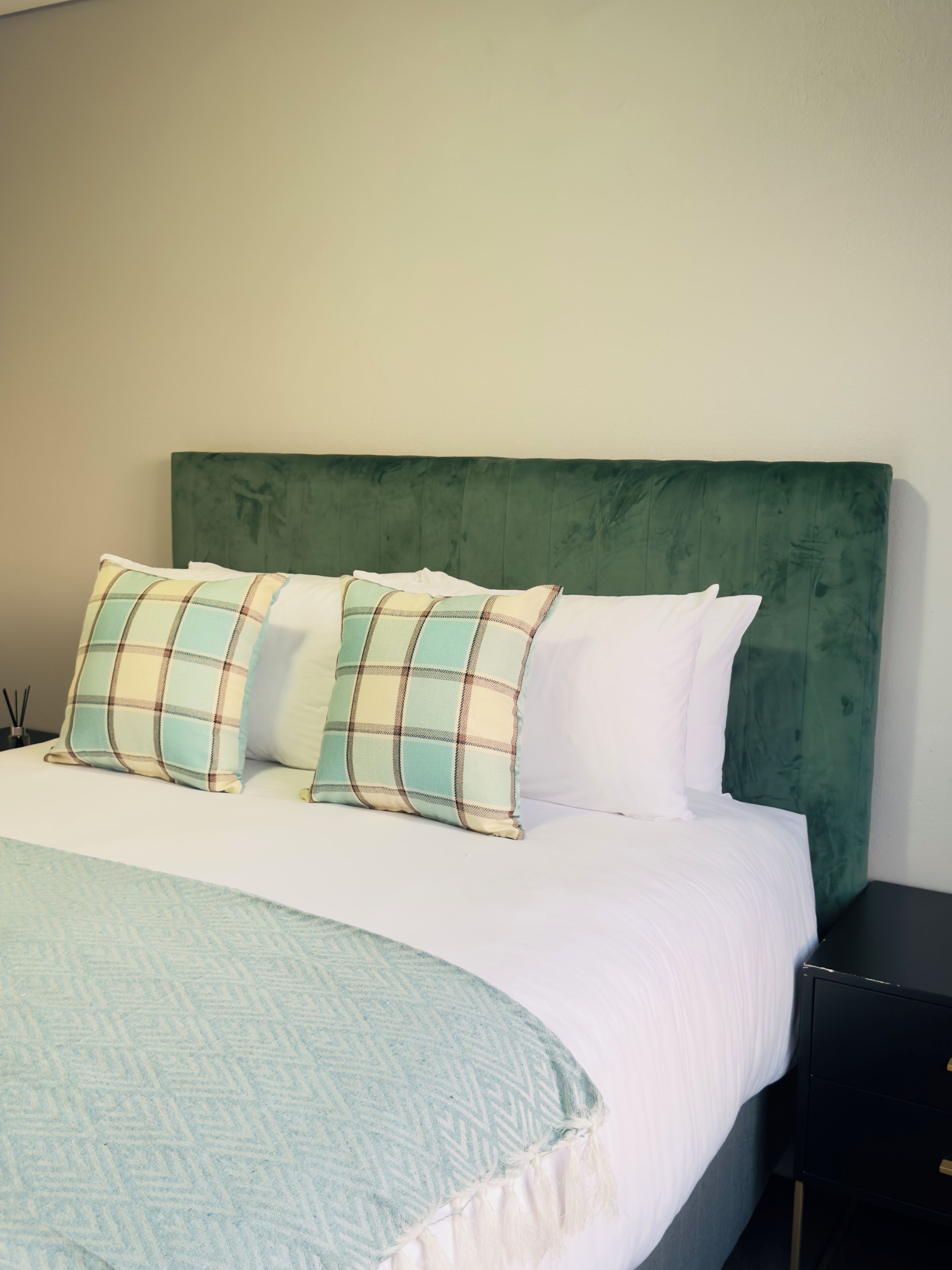A bed with a green upholstered headboard, white bedding, and two plaid pillows.