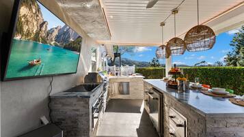 A modern outdoor kitchen with a large TV screen displaying a scenic view, a grill, and a dining area with a view of the mountains.