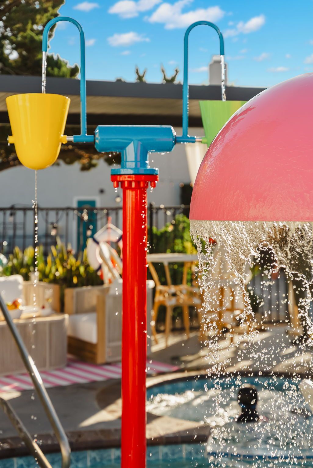 A water feature with a red spout and yellow and green containers, set against a poolside area with outdoor furniture.