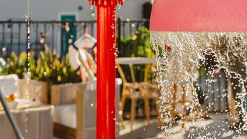 A water feature with a red spout and yellow and green containers, set against a poolside area with outdoor furniture.