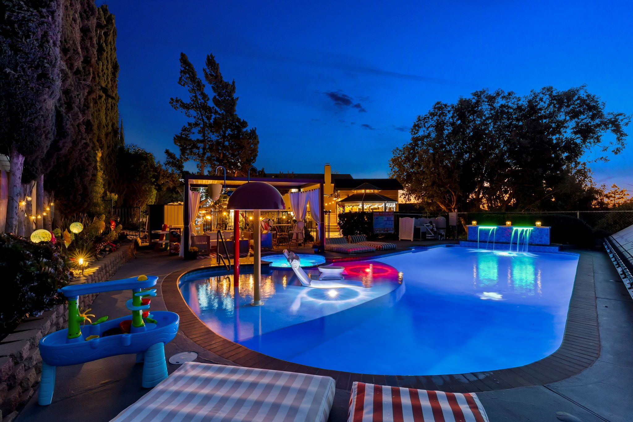 A swimming pool with a water feature, surrounded by lounge chairs and a covered seating area.