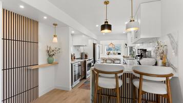 Una cocina moderna con armarios blancos, una isla central con taburetes y iluminación colgante, con vistas al exterior.