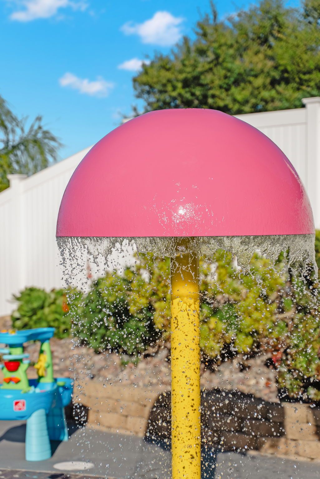 A water feature with a pink dome and a yellow pole, set against a backdrop of greenery and a white fence.