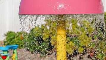 A water feature with a pink dome and a yellow pole, set against a backdrop of greenery and a white fence.