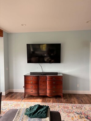 Ein Wohnzimmer mit einem an der Wand befestigten Flachbildfernseher, einem Holzschrank darunter und einem gemusterten Teppich auf dem Boden.