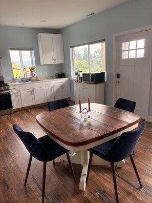 A dining area with a wooden table and chairs, a kitchen in the background, and a view of the outdoors. - Short Stop Stay in Homer. In private home cozy 2-bedroom apartment. (Homer)