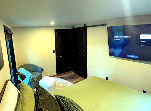 A hotel room with a bed, a seating area with a chair and cushions, a TV mounted on the wall, and a door leading to another room.