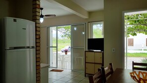 A kitchen with a refrigerator, a dining area with a table and chairs, a television on a stand, and a sliding glass door leading to an outdoor area with a car and greenery.
