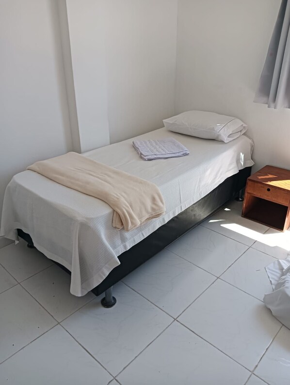 Ein Einzelbett mit weißer Matratze und einer beigen Decke, ein Nachttisch aus Holz und eine weiße Wand.