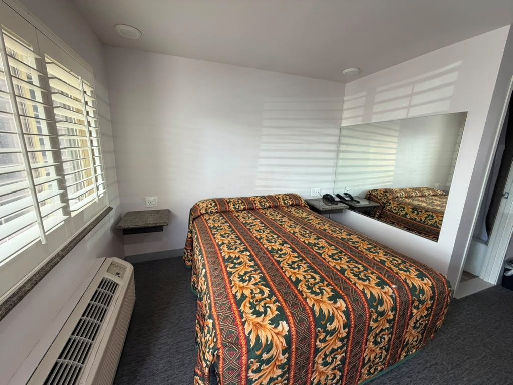 A hotel room with a bed, a mirror, a heater, and a window with blinds.