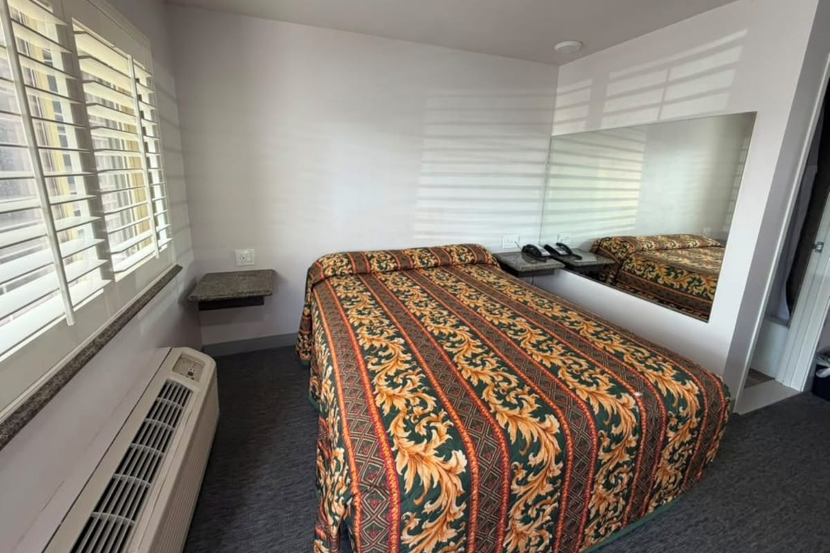 A hotel room with a bed, a mirror, a heater, and a window with blinds.