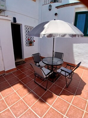 Un patio al aire libre con una mesa redonda y cuatro sillas, un paraguas, una pared blanca y una puerta.
