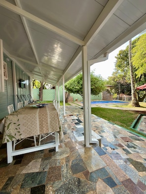 Un patio couvert avec une table à manger pour six personnes, une piscine extérieure et un sol carrelé en pierre.