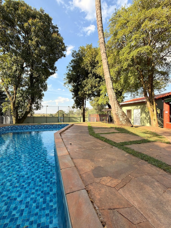 Une piscine dont le bord est revêtu de carreaux, entourée d’une terrasse en pierre et d’arbres.