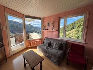 Quarto aconchegante com sofá, mesa pequena e cadeira vermelha. Possui janelas amplas com vista para um morro arborizado.