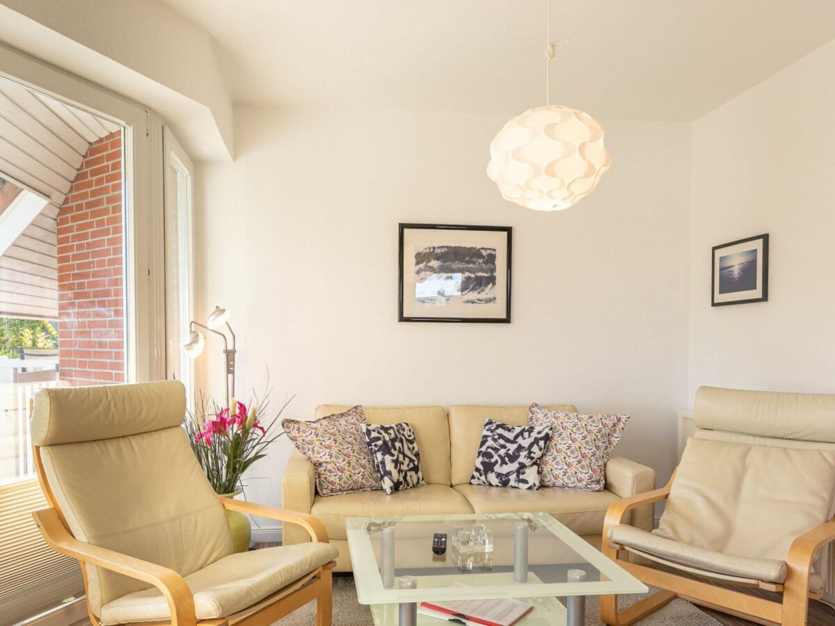 A living room with a sofa, two armchairs, a glass coffee table, a hanging light fixture, and two framed pictures on the wall.