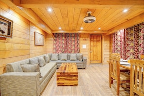 A cozy wooden interior with a sectional sofa, a wooden coffee table, and a dining area with a wooden table and chairs. - DanCenter Riverside Lexus Regency (Pahalgam)