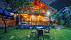 Uma cabana de madeira com varanda, base de pedra e um pequeno gramado com cadeiras.