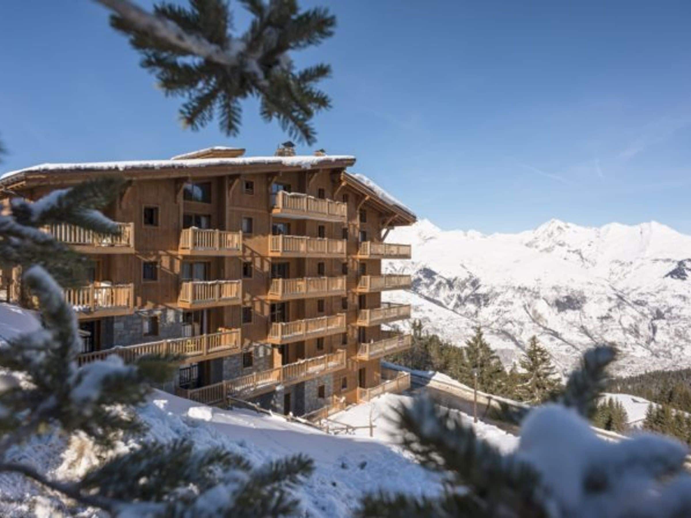 A multi-story wooden building with balconies, surrounded by snow-covered mountains and pine trees.