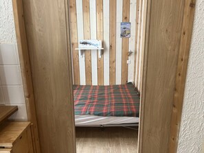 Quarto pequeno e estreito com parede de ripas de madeira, uma cama de solteiro com colcha xadrez, uma prateleira com adesivo de carro e uma porta branca.