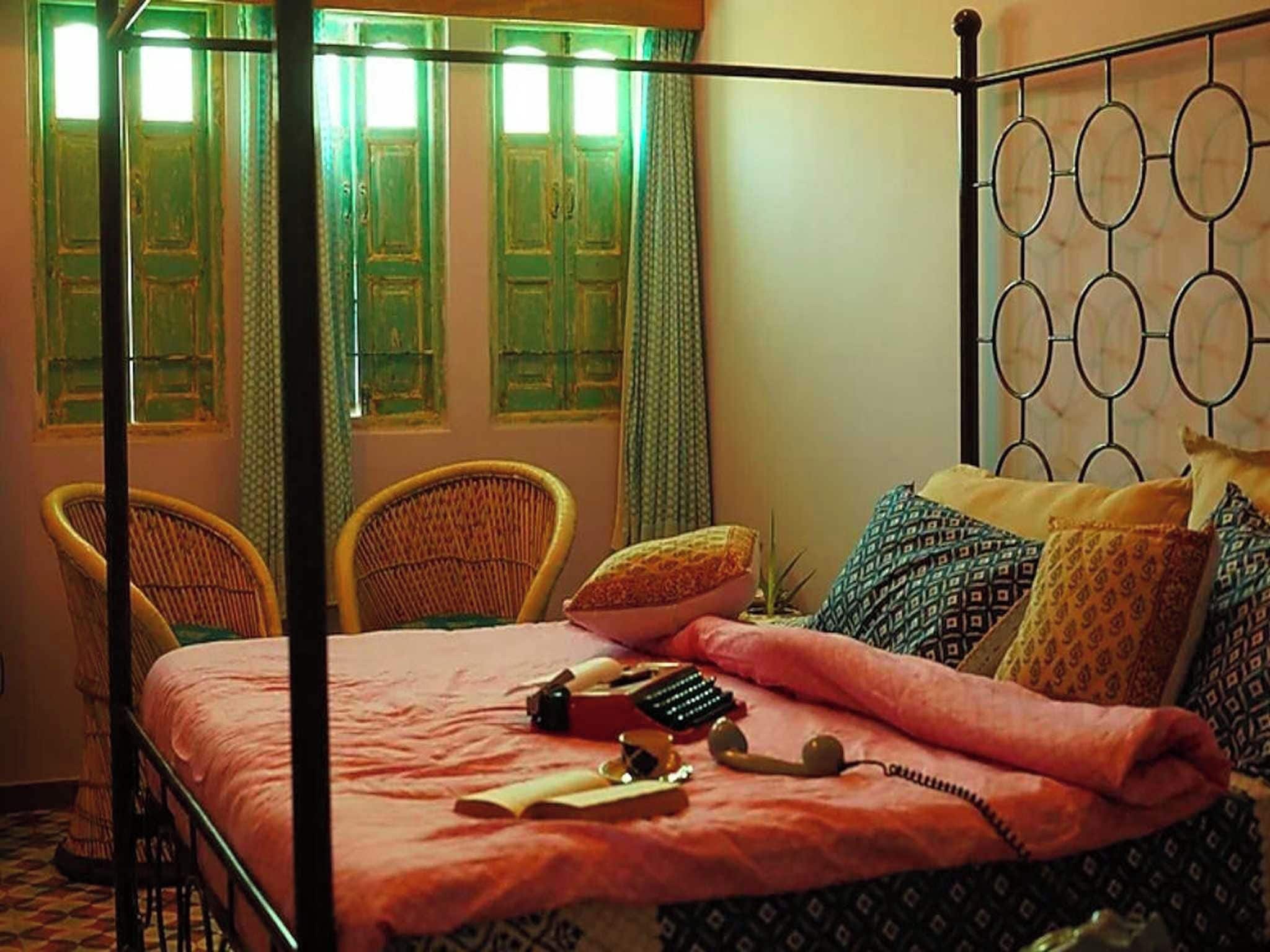 Una cama con dosel con colcha rosa, dos sillas de mimbre, cojines estampados y una mesita con teléfono y libros.