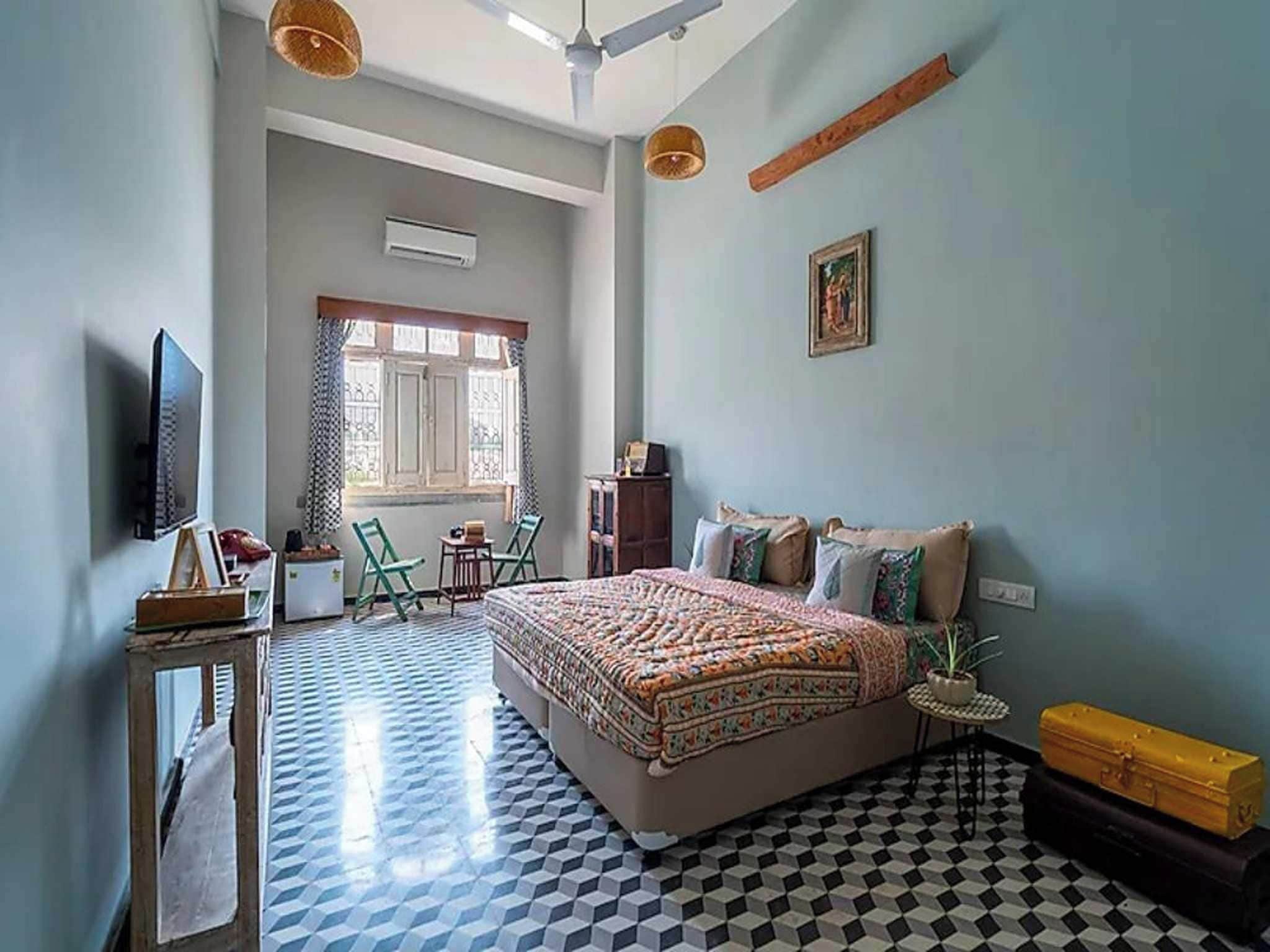 Un dormitorio con una cama grande, televisión, ventana con cortinas, una silla, una mesita y una maleta.