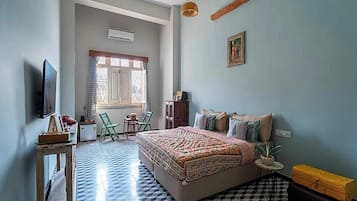 Un dormitorio con una cama grande, televisión, ventana con cortinas, una silla, una mesita y una maleta.