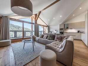 Ein modernes Wohnzimmer mit einem großen Sofa, einem runden Couchtisch und Blick auf eine Berglandschaft durch große Fenster.