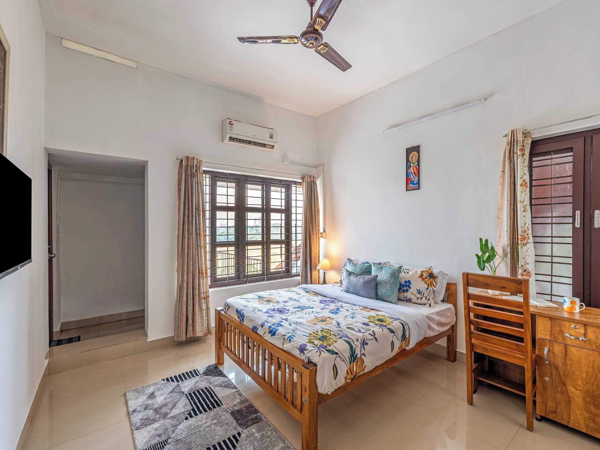 Un dormitorio con cama, escritorio, silla, ventilador de techo, ventana con cortinas, equipo de aire acondicionado y puerta.