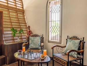 A room with a chessboard on a table, two chairs with cushions, a window with a metal grill, and a plant on the windowsill.