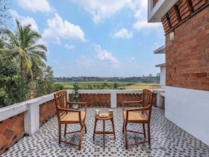 A balcony with two wooden chairs and a small table, a potted plant, and a view of a cityscape and greenery.