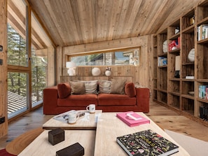 Een gezellige kamer met een houten plafond, een rode bank, een bijzettafeltje met boeken en een mok, en een boekenplank vol boeken en decoratieve objecten.