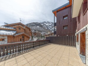 A building with a wooden facade and a stone base, a metal fence, and a mountainous backdrop. - Charming apartment - 7 guests, south terrace (Val-d'Isère)
