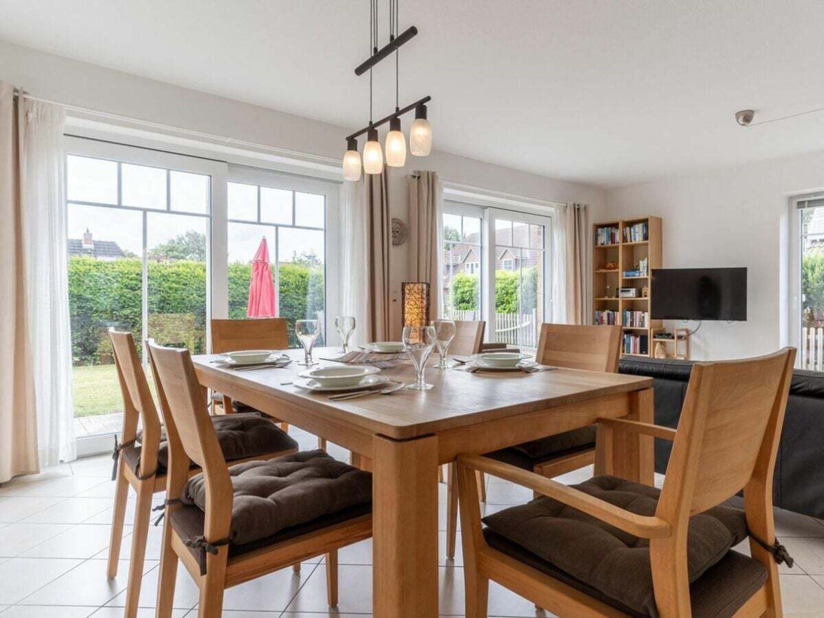 Un comedor con mesa y sillas de madera, una araña y vistas al exterior a través de amplios ventanales.