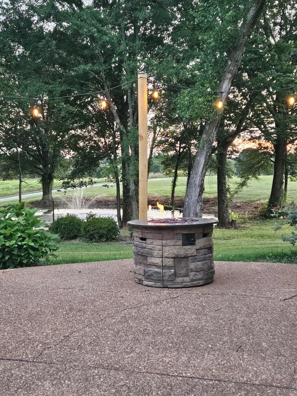 A stone fire pit with a wooden post in a landscaped area with trees and a grassy field.