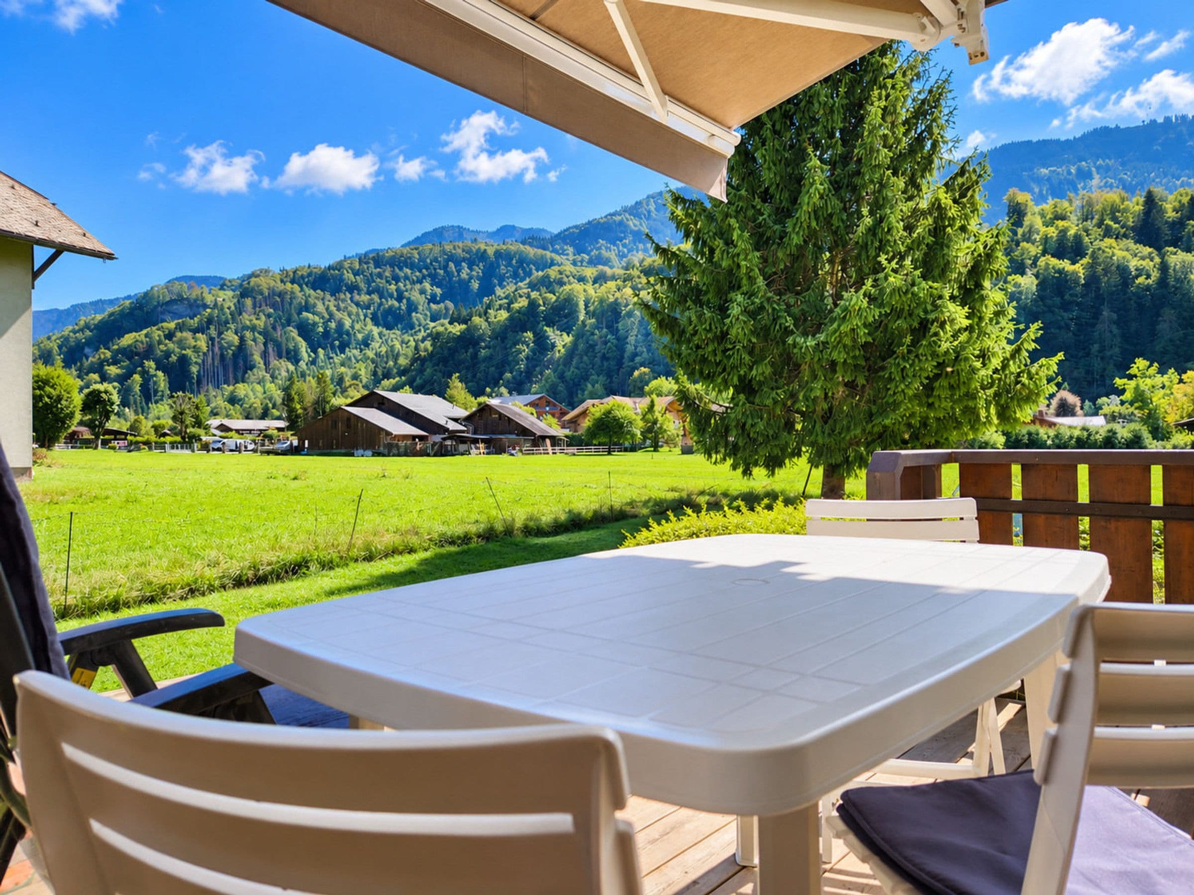 A patio with a table and chairs, overlooking a green field and a mountain range.