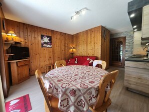 A cozy room with wooden walls, a round table with a patterned tablecloth, a red sofa, and a television on a wooden cabinet. - Ideal T2 apartment for 4 guests - Samoëns (Samoëns)
