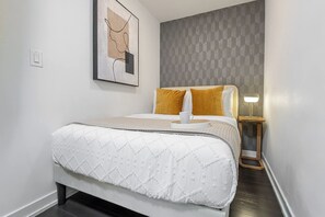 Una cama bien hecha con colcha blanca, una mesita de noche con lámpara y una pared con diseño geométrico.