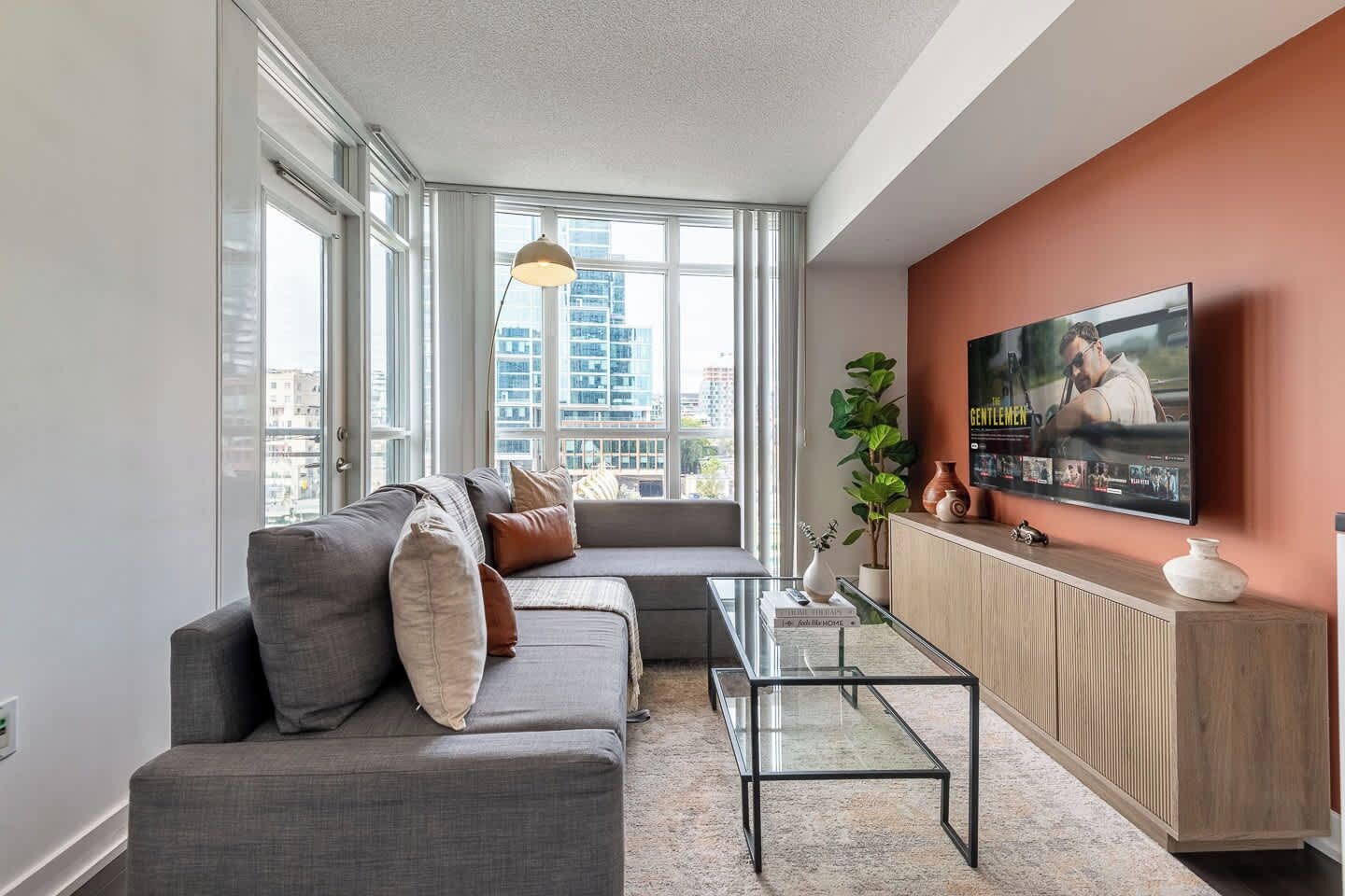Een moderne woonkamer met een grijze modulaire bank, een glazen salontafel, een aan de muur bevestigde televisie en een groot raam met uitzicht op de stad.