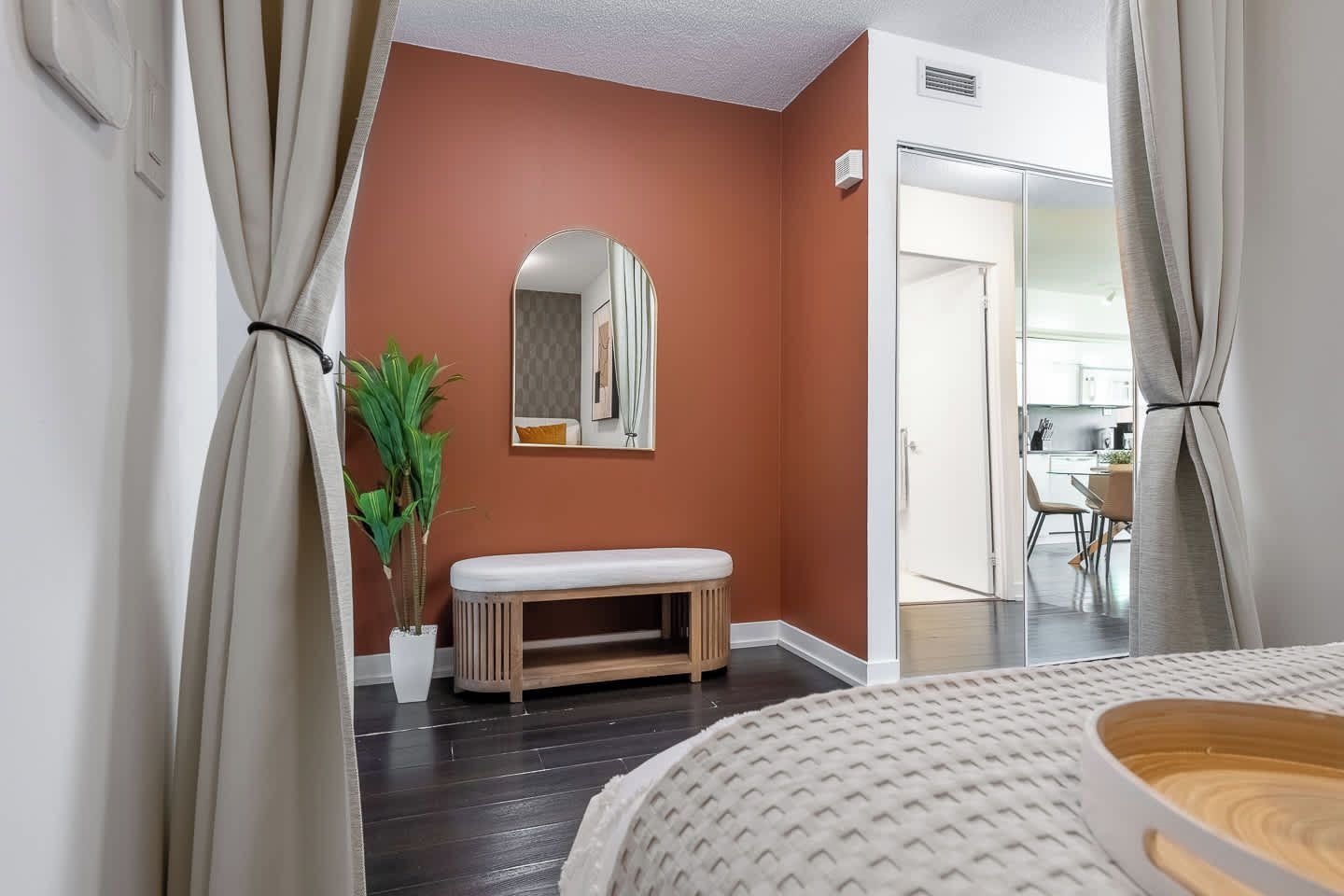 A hotel room with a wooden bench, a potted plant, a mirror on the wall, and a view of the kitchen through an open door.