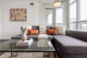 Una sala moderna con una mesa de centro de vidrio, un sofá gris con múltiples cojines, un florero con una planta y vistas a la ciudad a través de ventanas amplias.